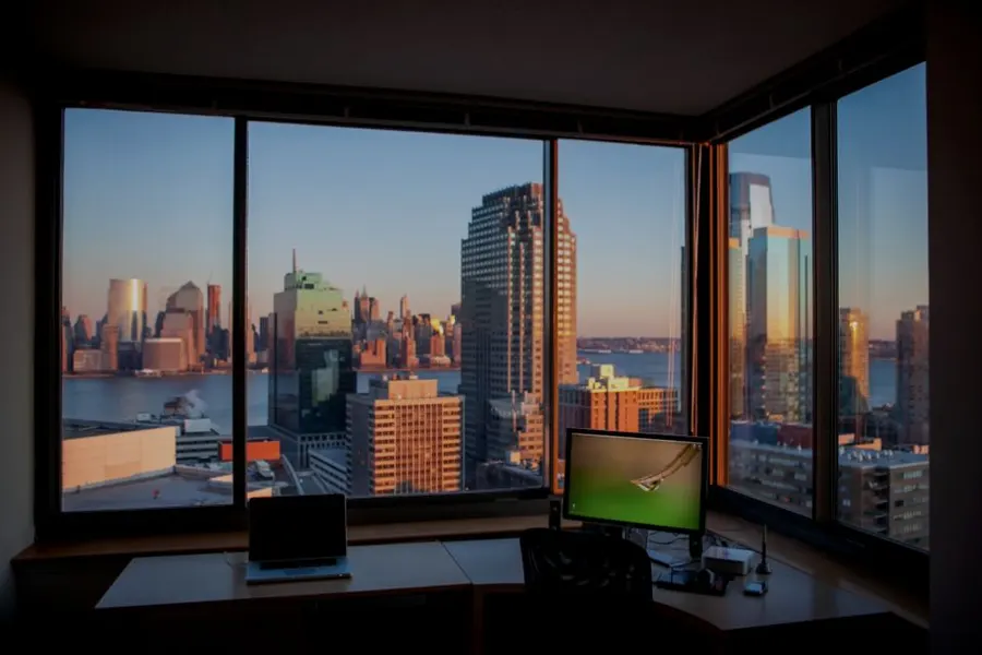 A desk with a laptop and monitor overlooking a city skyline at dusk